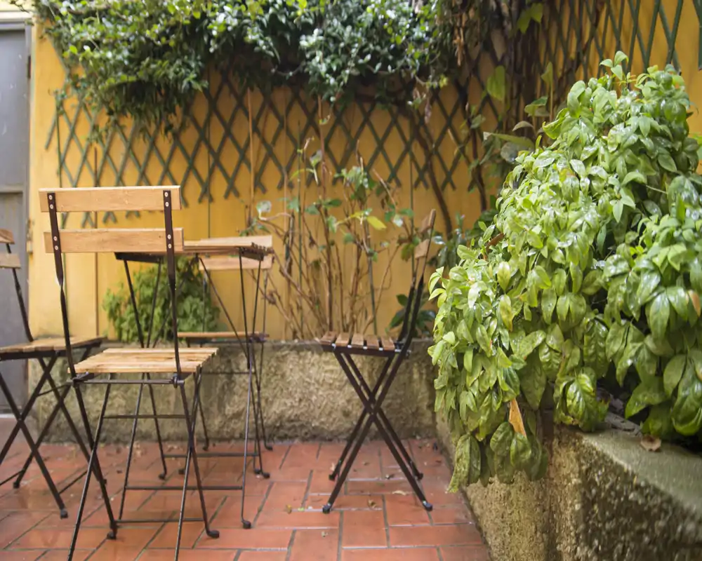 Acogedora terraza con suelo de baldosas, sillas plegables de madera y mesa, enmarcada por plantas verdes y una pared amarilla cálida — ideal para un café al aire libre.