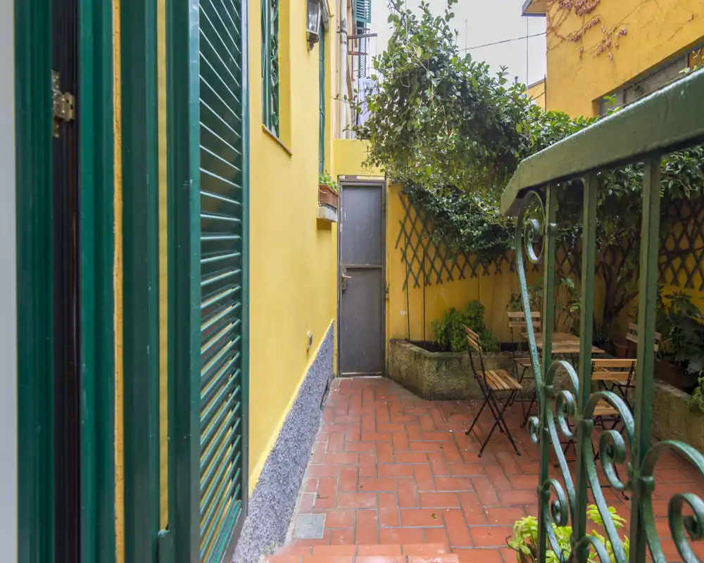 Pequeño patio privado con suelo de terracota, contraventanas verdes y una mesa con sillas plegables bajo plantas trepadoras: una tranquila zona exterior.