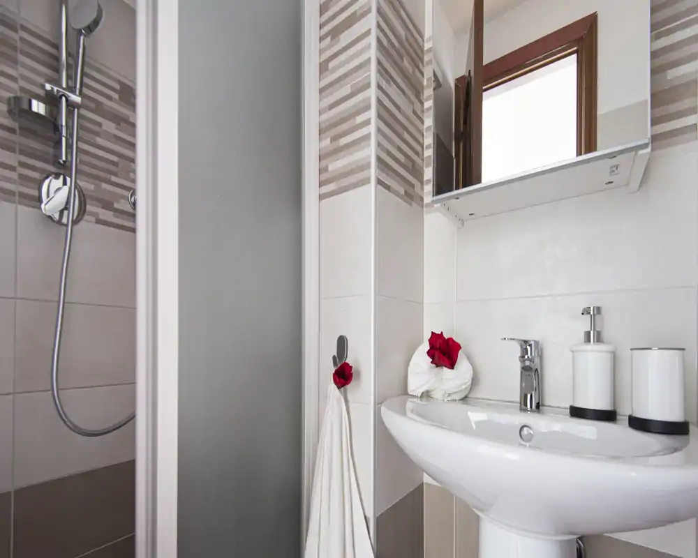 Baño compacto y limpio con lavabo pedestal, grifo cromado y ducha con azulejos. Toallas frescas y pequeños detalles rojos decorativos aportan una sensación ordenada y acogedora.