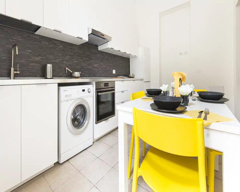 Cocina moderna y luminosa con electrodomésticos integrados y una pequeña mesa puesta; las sillas amarillas aportan un toque alegre.