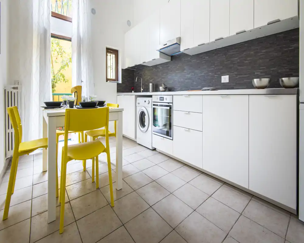 Cocina moderna y luminosa con armarios blancos, electrodomésticos integrados y una mesa pequeña con sillas amarillas alegres — ideal para un piso compacto.
