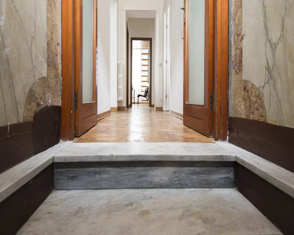 Entrada luminosa con escalones de mármol que conducen a un pasillo con parquet de madera y puertas dobles de madera; la vista continúa hasta una habitación amueblada.