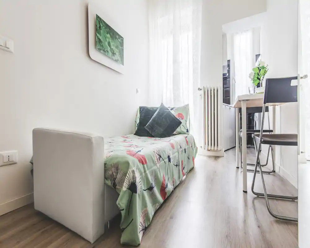 Compact, bright bedroom with a single bed dressed in patterned linens, soft cushions and natural light from tall windows — ideal for a cozy city stay.