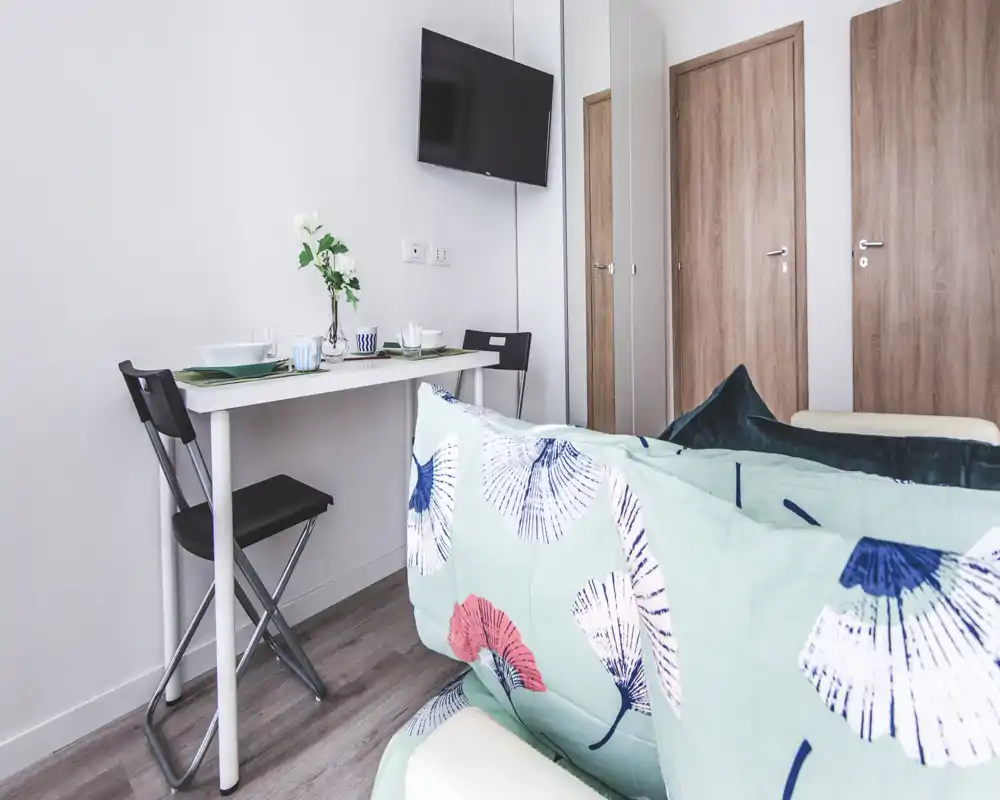 Compact living area with a small dining/desk table, wall-mounted TV and a sofa with patterned cushions — bright, modern and functional for short stays.
