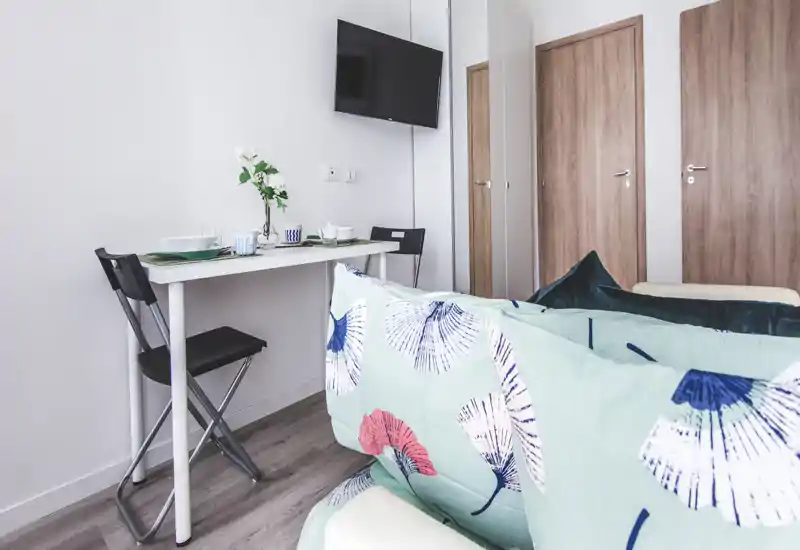 Compact living area with a small dining/desk table, wall-mounted TV and a sofa with patterned cushions — bright, modern and functional for short stays.