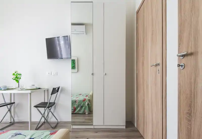 Bright, minimal bedroom corner with a wardrobe featuring a full-height mirror, a wall-mounted TV, and a glimpse of a bed with a floral bedspread reflected in the mirror. Simple dining set and light wood doors complete the functional layout.