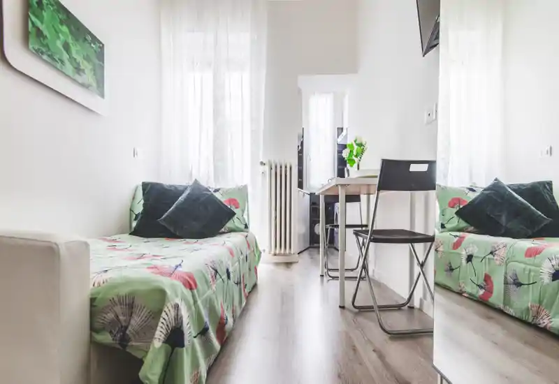 Bright, compact bedroom with two single beds/sofa-beds dressed in patterned green covers, dark cushions and sheer curtains letting in natural light; small desk and chair create a functional, cozy layout ideal for a short-stay rental.