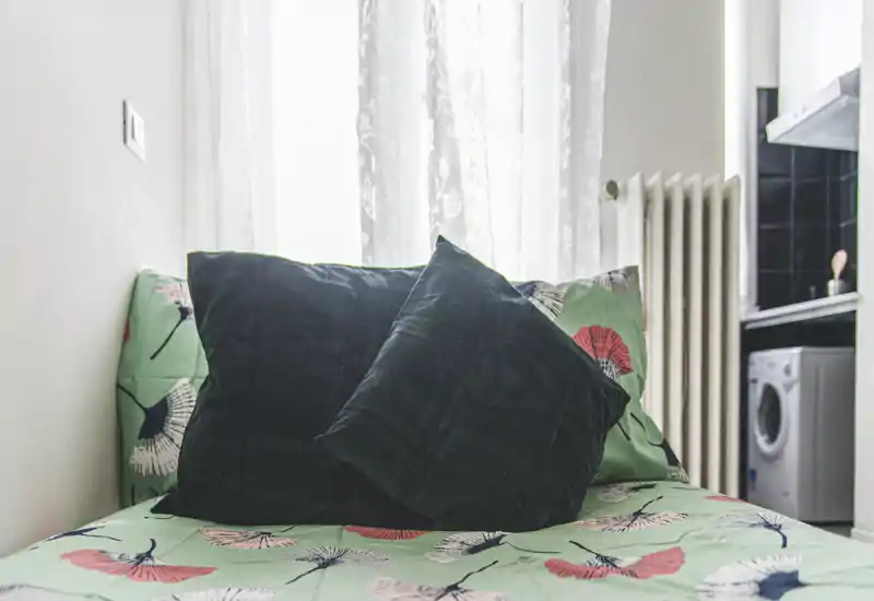 Bright, cozy bedroom corner with patterned bedding and plush dark green pillows placed against a sheer-curtained window.