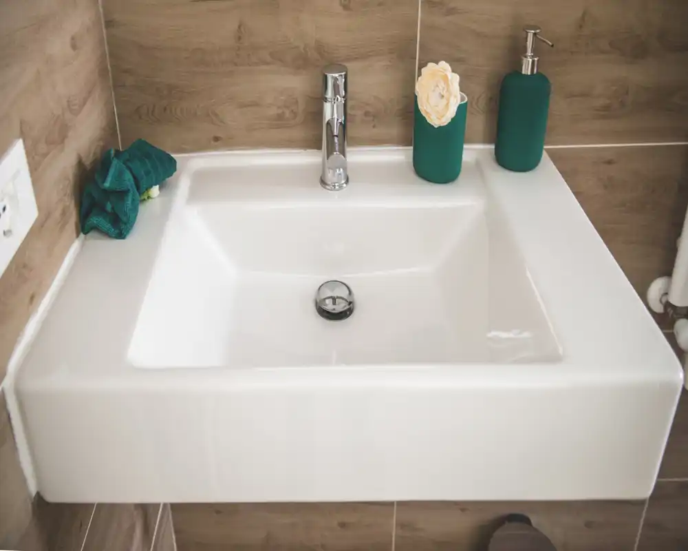 Close-up of a modern white bathroom sink with chrome faucet and green accessories on a wood-effect tiled wall.