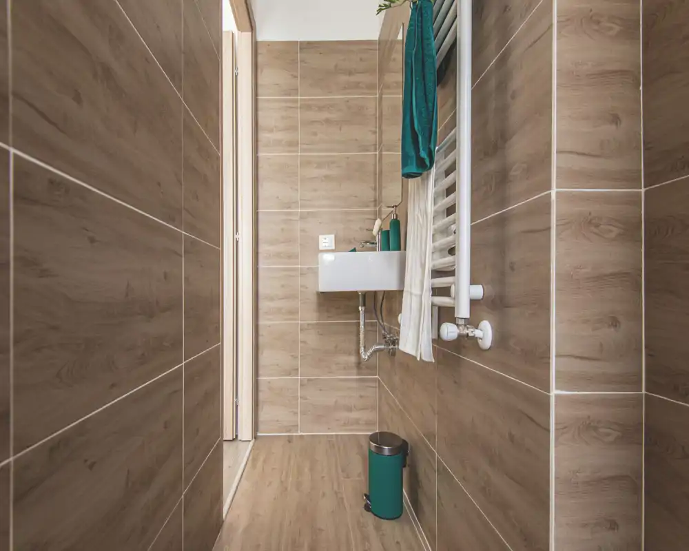 Narrow modern bathroom with wood-effect tiles, a wall-mounted sink, towel radiator and coordinated teal accessories.