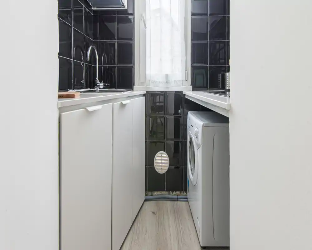 Compact kitchen/laundry corner with white cabinets, black glossy tiled backsplash, a sink and a front-loading washing machine under the counter; bright window provides natural light.