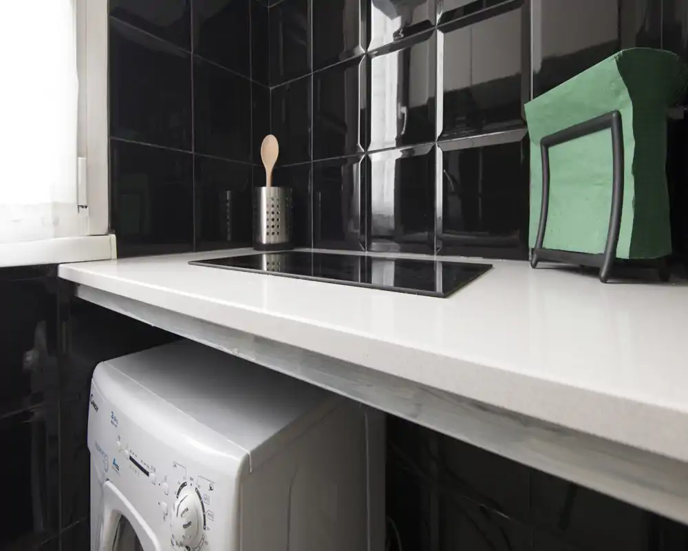 Compact modern kitchen corner with a built-in electric cooktop, glossy black tiled backsplash and a small appliance (washing machine) under the counter.