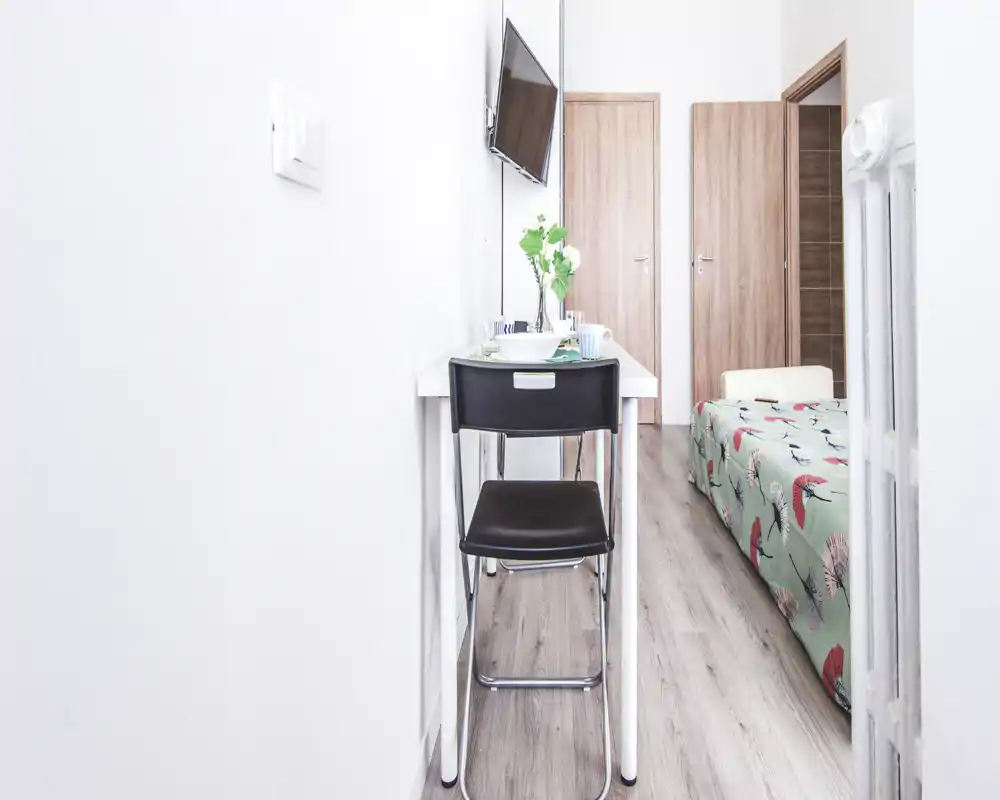Compact, bright bedroom corner with a single bed dressed in a colorful patterned cover and a narrow desk with chair and wall-mounted TV. Fresh, minimal styling and wood-effect floors give a neat, welcoming feel.
