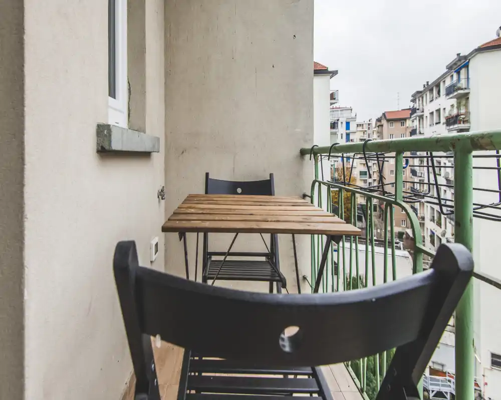 Small furnished balcony with a wooden table and folding chairs, overlooking neighboring apartment buildings — ideal for morning coffee.