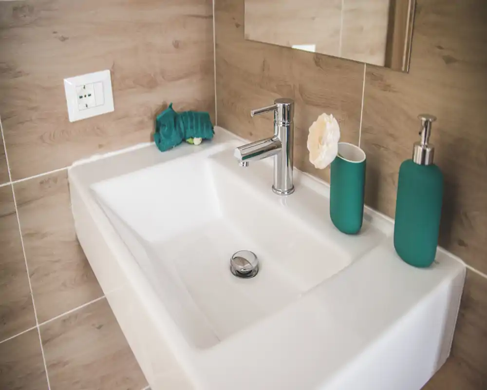 Close-up of a modern bathroom sink with chrome faucet, soap dispenser and matching cup, warm wood-effect tiles and a mirror reflecting part of the wall.