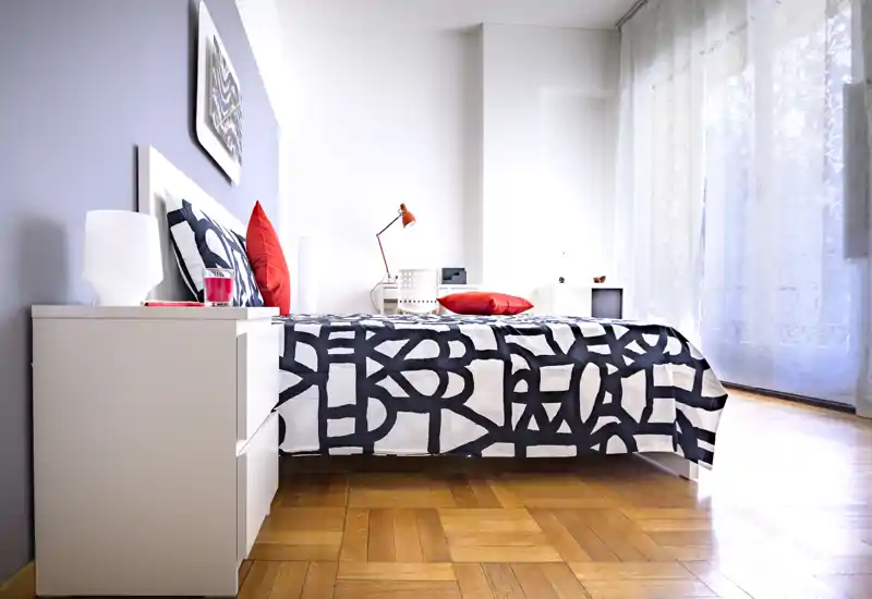 Chambre moderne et lumineuse avec couette à motifs, coussins rouges en accent et grande fenêtre apportant une belle lumière naturelle. Table de chevet épurée et parquet chaud pour une ambiance contemporaine idéal pour une annonce.