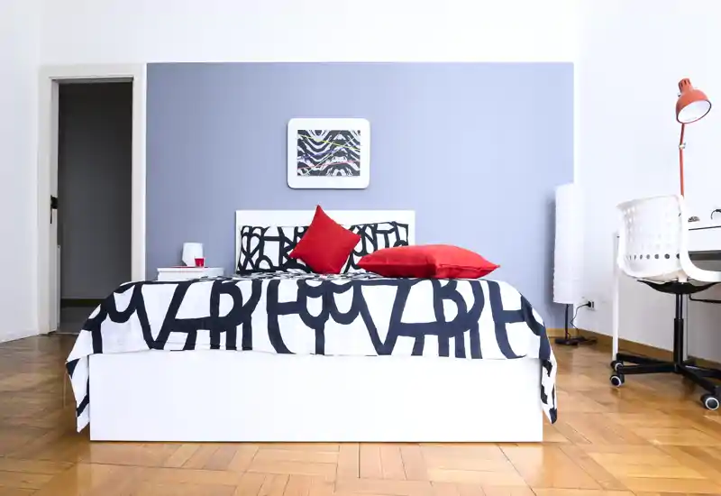 Chambre moderne et lumineuse avec un lit double centré, literie à motifs audacieux et coussins rouges. La pièce comprend un bureau soigné, un sol en parquet et une lumière naturelle douce.