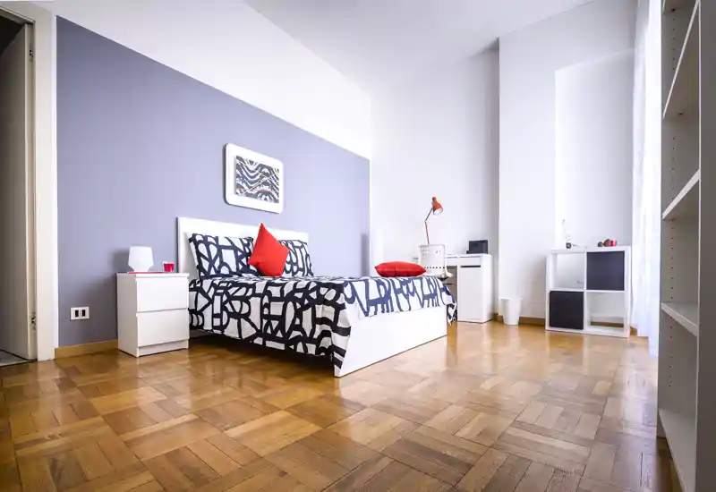 Chambre lumineuse et stylée avec lit complet, linge de lit à motifs audacieux et coussins rouges; grande fenêtre apportant lumière naturelle et mobilier moderne épuré.