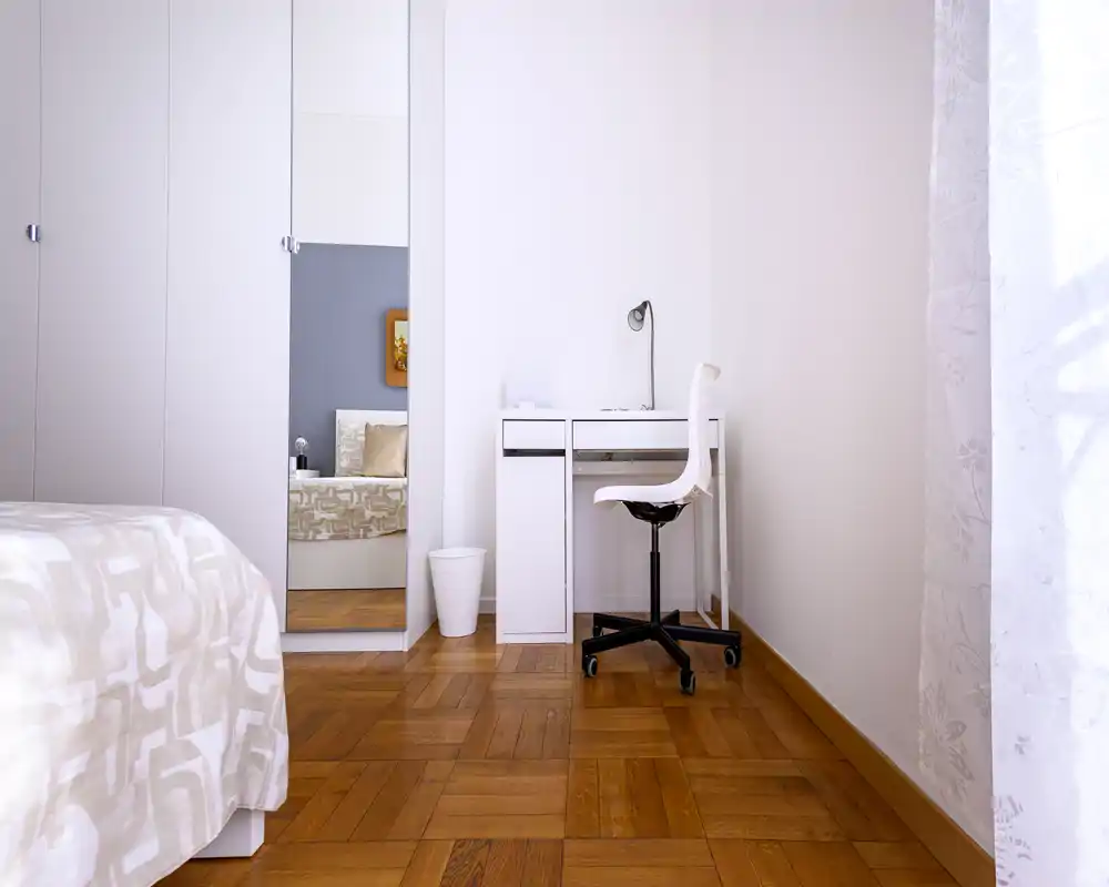 Bright, minimal bedroom with a white wardrobe, a visible bed with patterned bedding, and a small white desk by the window — clean lines and warm wooden flooring create a calm atmosphere.