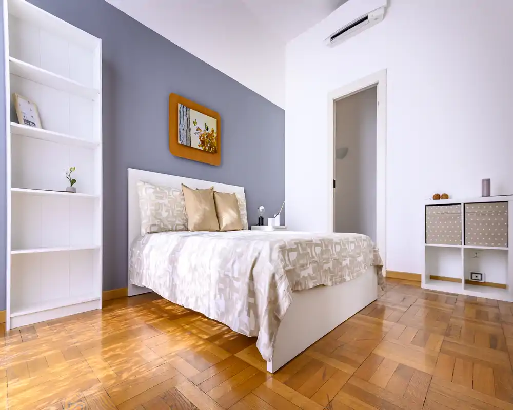 Bright, minimal bedroom with a neatly made double bed, decorative pillows, wooden parquet flooring and simple shelving — a calm, well-lit space ideal for rentals.