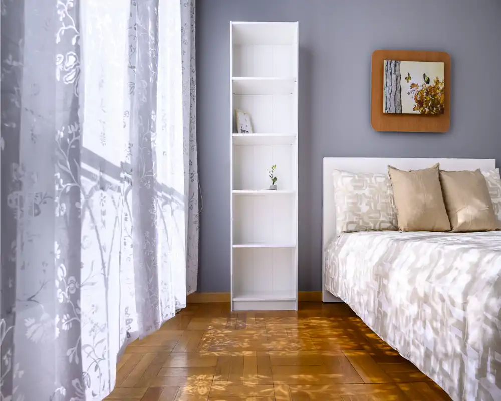 Bright, cozy bedroom with a neatly made bed, warm wood flooring and soft curtains filtering natural light.