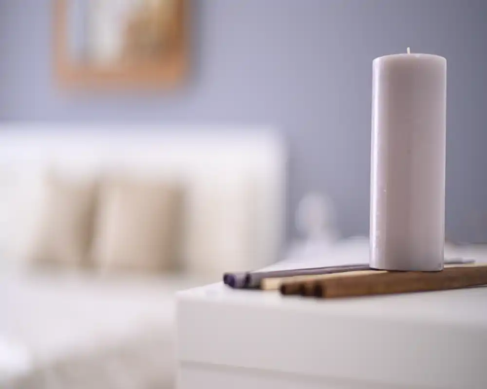 A softly lit bedroom detail with a decorative candle in the foreground and a blurred headboard and pillows in the background, creating a calm, cozy atmosphere.