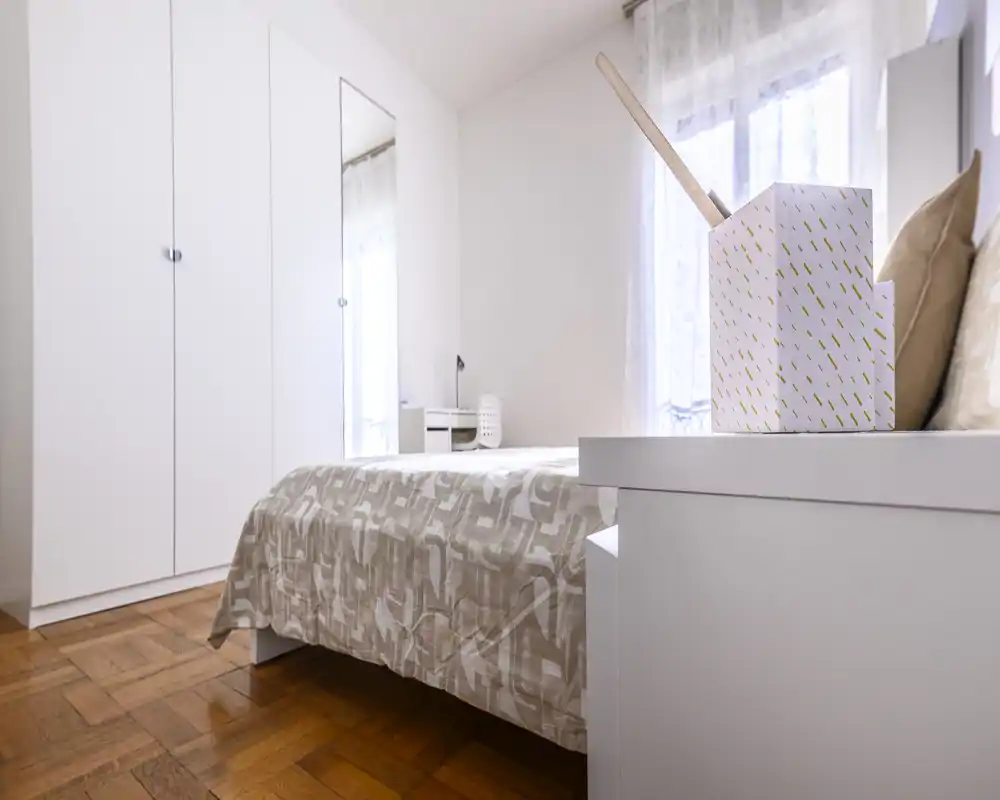 Bright, minimal bedroom with a neatly made bed, white wardrobe and natural light from a window. The neutral textiles and wooden parquet floor give a warm, calm atmosphere ideal for rental listings.