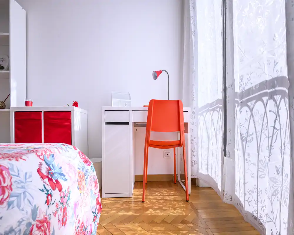 Rincón de dormitorio luminoso y alegre con ropa de cama floral, un escritorio blanco y una silla naranja junto a la ventana soleada.