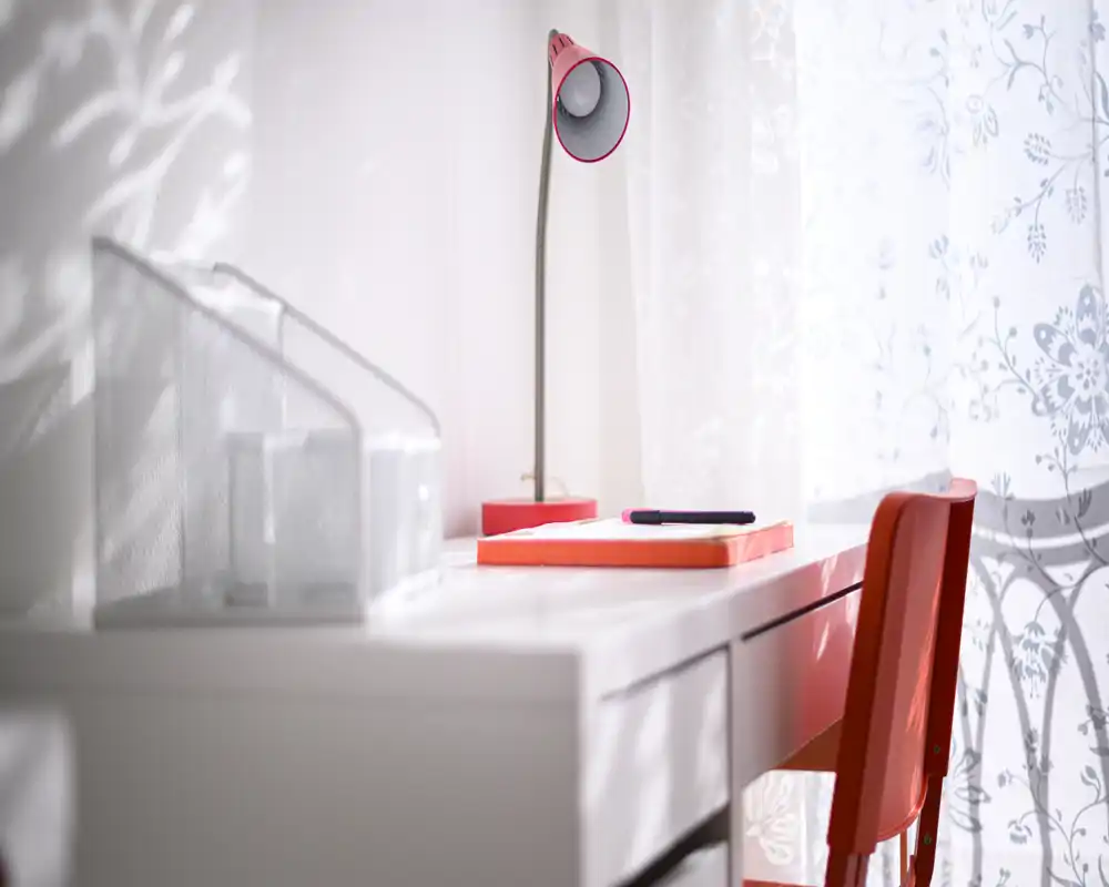 Rincón de trabajo luminoso y bien decorado con escritorio blanco, silla roja y lámpara a juego junto a una cortina ligera estampada — ideal para un pequeño despacho en casa.