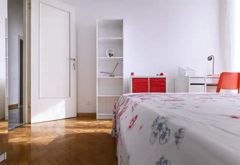 Camera da letto luminosa e moderna con biancheria floreale in primo piano, una libreria bianca e una piccola scrivania con sedia arancione vicino alla finestra. La luce naturale e il pavimento in parquet donano un’atmosfera accogliente.