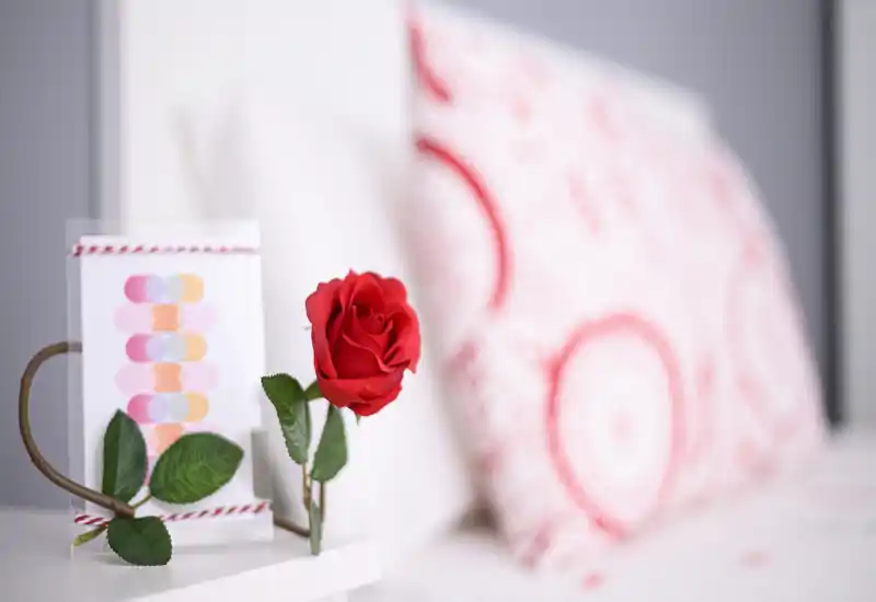 Primo piano del comodino della camera da letto con una rosa rossa e un cuscino decorato in un ambiente luminoso e accogliente — perfetto per mostrare dettagli decorativi.