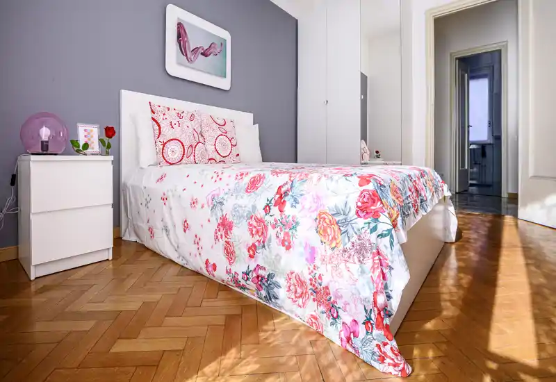 Camera da letto luminosa e ben arredata con copriletto floreale, cuscini decorativi e pavimento in parquet illuminato dal sole — accogliente e curata.