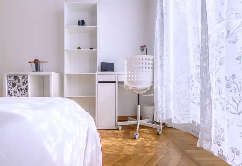 Rincón de dormitorio luminoso y aireado con cama visible, escritorio blanco y silla junto a cortinas translúcidas con motivos florales que dejan pasar la luz natural.