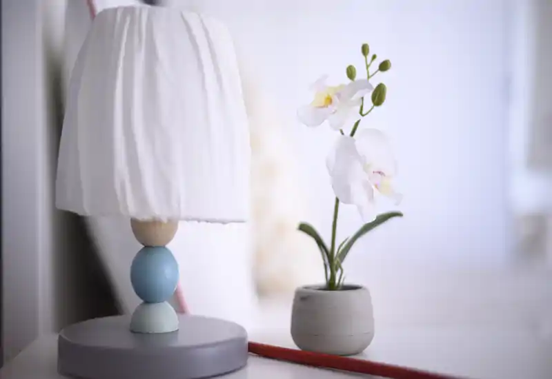 Primer plano de una mesita con una lámpara decorativa y una pequeña orquídea, fotografiado con luz suave y luminosa — ideal para destacar detalles de estilo en el dormitorio.