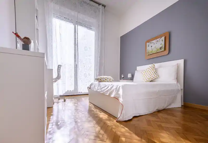 Dormitorio moderno y luminoso con cama blanca, cojines estampados, lámpara de noche y gran ventana con cortinas traslúcidas que dejan pasar mucha luz natural. Suelo de parquet cálido y decoración minimalista crean un ambiente acogedor ideal para anuncios.