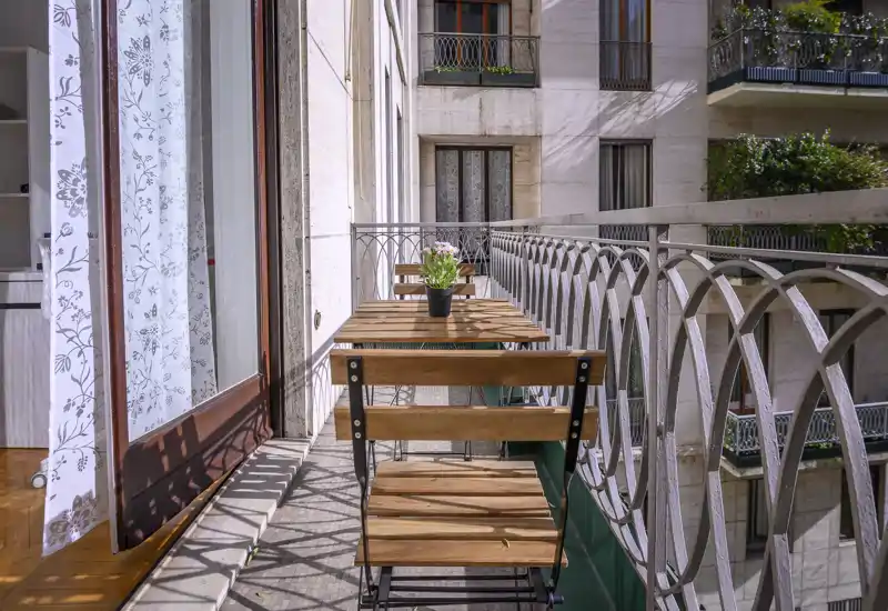 Balcón soleado y estrecho con varias mesas y sillas bistró de madera, barandilla decorativa y una pequeña maceta — perfecto para el café de la mañana.