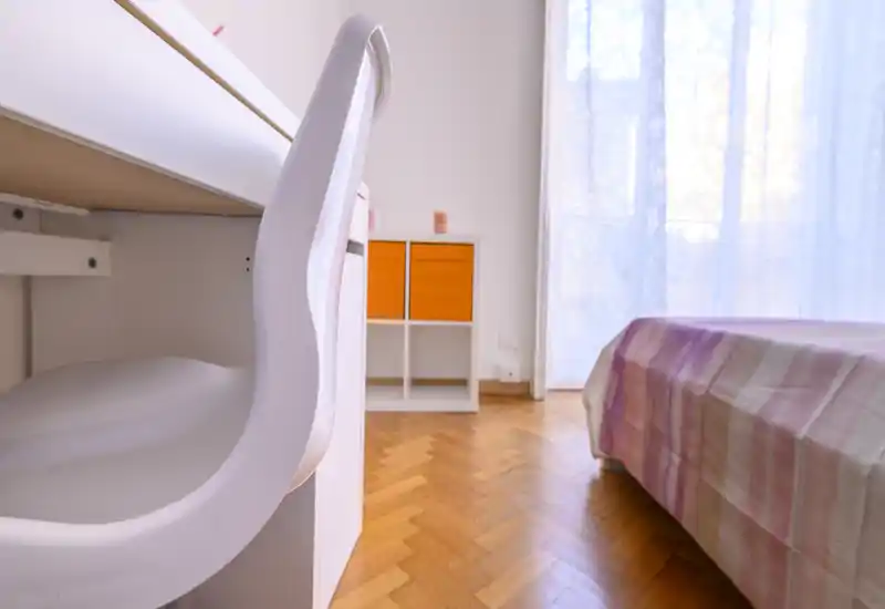 Rincón de dormitorio luminoso y acogedor con cama visible con ropa de cama en tonos pastel, suelo de parquet y luz natural de una gran ventana — ideal para una estancia cómoda.