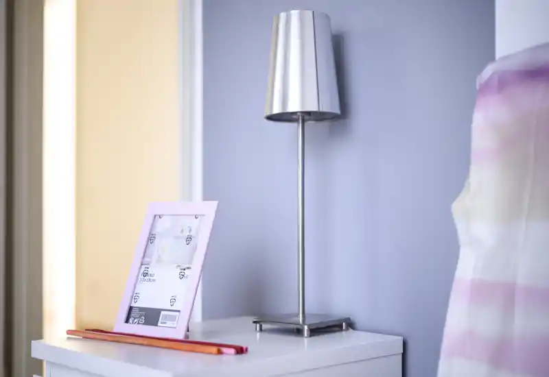 Rincón de dormitorio luminoso con mesita blanca, una lámpara metálica delgada y un portafotos; se ve parte de la cama con ropa de cama a rayas en el borde derecho.
