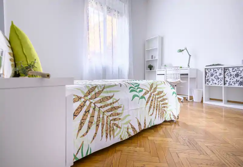 Dormitorio luminoso y moderno con colcha estampada, muebles blancos, escritorio y luz natural desde la ventana — espacio limpio y acogedor, ideal para una persona o estudiante.