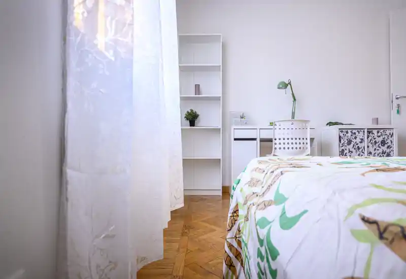 Dormitorio luminoso y minimalista con edredón estampado, escritorio y estanterías; la luz natural atraviesa cortinas translúcidas sobre el cálido suelo de parquet.