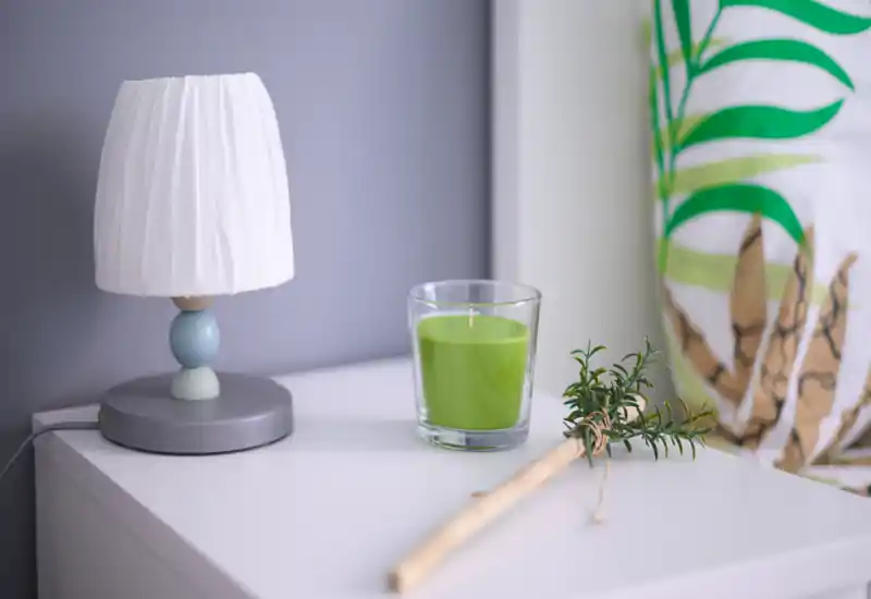 Primer plano de una mesita de noche con una lámpara pequeña, una vela verde en vaso y una rama decorativa, detalle de dormitorio estilizado.