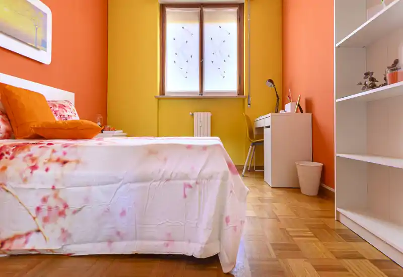 Camera da letto luminosa e accogliente con pareti arancioni e gialle, copriletto floreale e una scrivania vicino alla finestra — arredo moderno e ordinato, ideale per una singola persona.