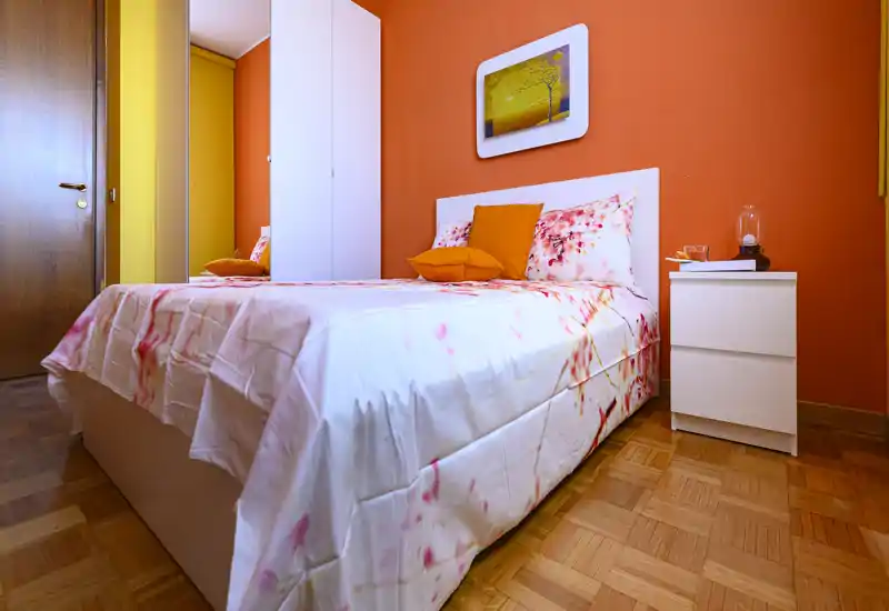 Camera da letto luminosa e calda con letto bianco, biancheria floreale e cuscini arancioni. Presenti un armadio bianco, un comodino e pareti arancioni che rendono l'ambiente accogliente e moderno.