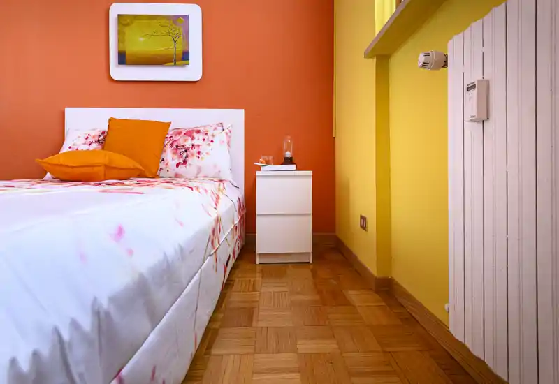 Camera da letto luminosa e accogliente con parete arancione, biancheria a fantasia e un piccolo comodino bianco; pavimento in legno e cuscini vivaci creano un ambiente invitante.