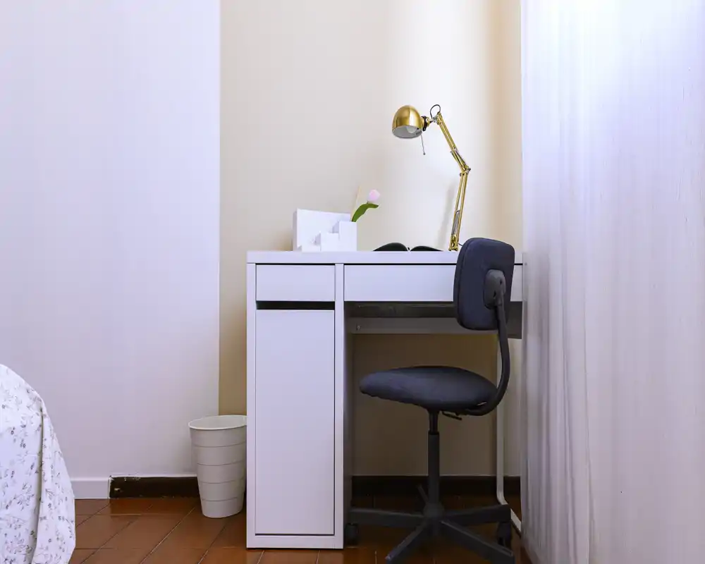 Angolo camera con una scrivania bianca compatta, lampada da tavolo e sedia girevole accanto al bordo del letto parzialmente visibile — luminoso e ordinato, adatto a una persona.