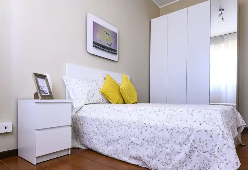 Camera da letto luminosa e ordinata con copriletto a fantasia, cuscini gialli come accento e un grande armadio bianco—perfetta per annunci che evidenziano comfort e spazio di stivaggio.