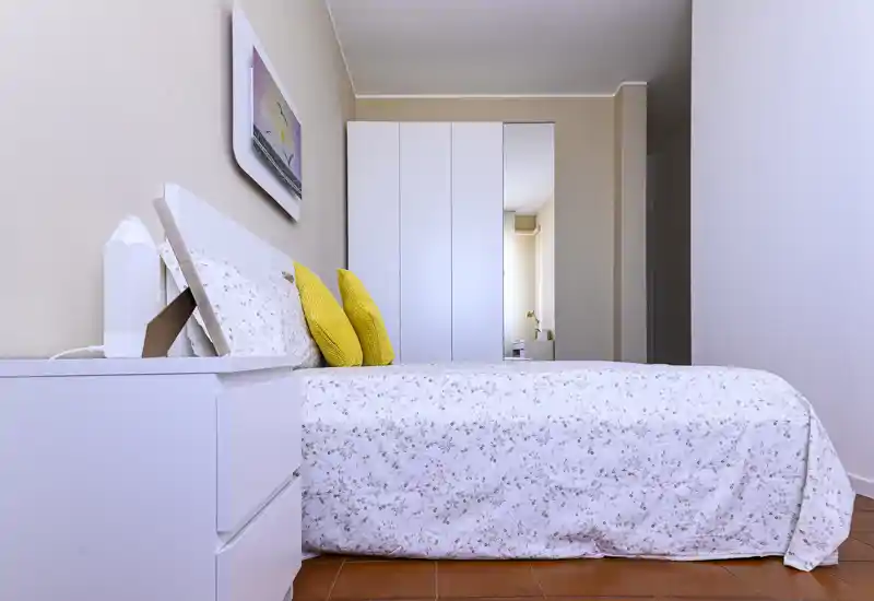 Camera da letto luminosa e ordinata con letto matrimoniale con lenzuola bianche stampate e cuscini gialli; armadio a muro con anta specchiata che aggiunge profondità e spazio.