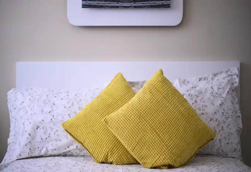 Camera da letto luminosa e accogliente in primo piano: letto rifatto con lenzuola fantasia e due cuscini decorativi gialli a trama, appoggiati su una testiera bianca semplice.