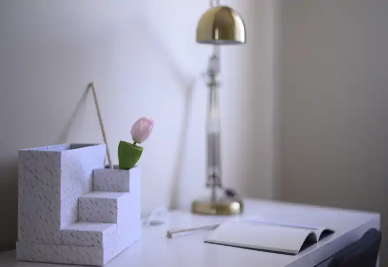 Un coin bureau soigné, type petit espace salon/travail : rangement décoratif avec une tulipe, une lampe de bureau en laiton et un carnet ouvert sur une surface blanche.