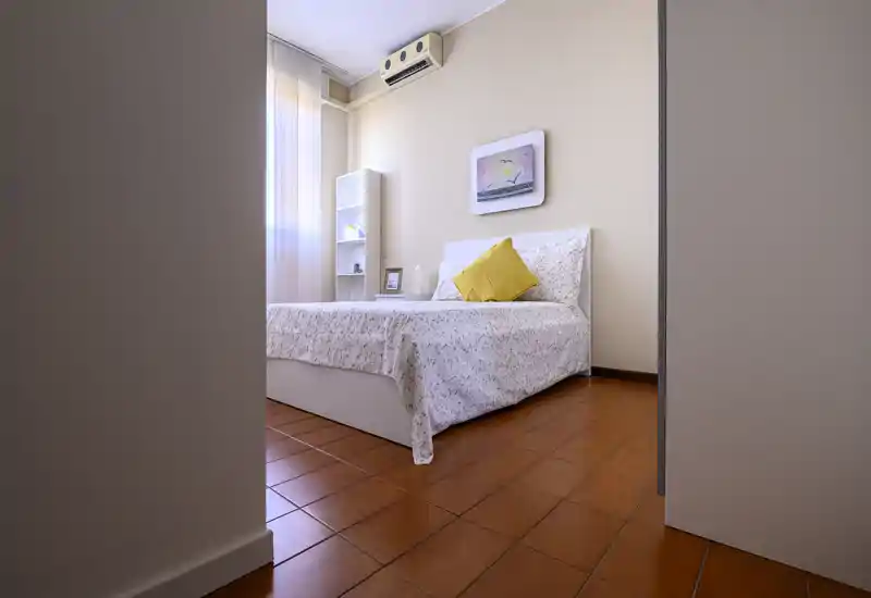 Camera da letto ordinata e ben illuminata con letto matrimoniale bianco, biancheria a fantasia e cuscino giallo a contrasto; una libreria alta e un quadro completano l’arredo sobrio.